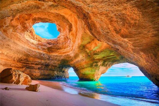 Grotte marine de l'Algarve aux parois orange, ouverte sur une plage de sable et une mer turquoise sous un ciel bleu lumineux.
