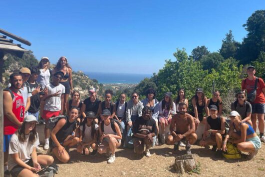 Sous un ciel bleu, un groupe Eole Loisirs pose lors d’une excursion en Crète, avec la mer en arrière-plan et une ambiance conviviale.